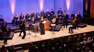 Ein klassisches Musikensemble spielt auf der Bühne mit Streich- und Blasinstrumenten. Eine Frau in einem schwarzen Kleid steht vor einem Pult. Das Publikum sitzt im Vordergrund.