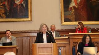Eine Frau spricht an einem Podium, mit einem Mikrofon vor ihr. Hinter ihr sitzt ein Mann an einem Schreibtisch, der einen Anzug und eine Krawatte trägt. Zwei große gerahmte Gemälde schmücken die Wand.