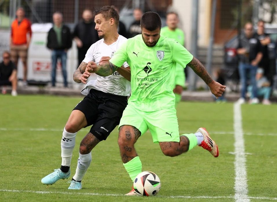 Zwei Fußballspieler in Aktion, einer in Grün, der andere in Weiß, kämpfen um den Ball auf einem grünen Feld mit Zuschauern im Hintergrund.
