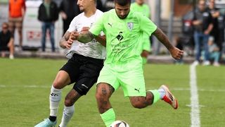 Zwei Fußballspieler in Aktion, einer in Grün, der andere in Weiß, kämpfen um den Ball auf einem grünen Feld mit Zuschauern im Hintergrund.