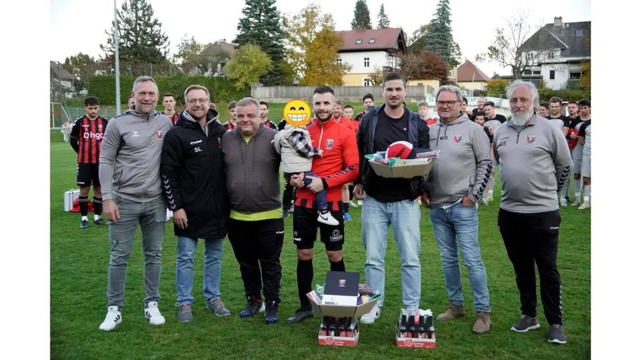 Eine Gruppe von Männern steht auf einem Feld mit einem Kind, das einen Fußball hält, während andere Geschenke halten und lächeln. Bäume und Häuser sind im Hintergrund.