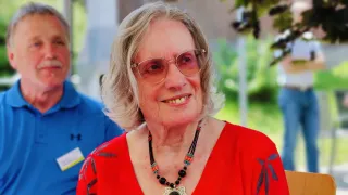 Eine ältere Frau mit weißem Haar, die ein rotes Shirt und eine Halskette trägt, lächelt für die Kamera. Sie trägt eine Brille und scheint sich im Freien aufzuhalten.