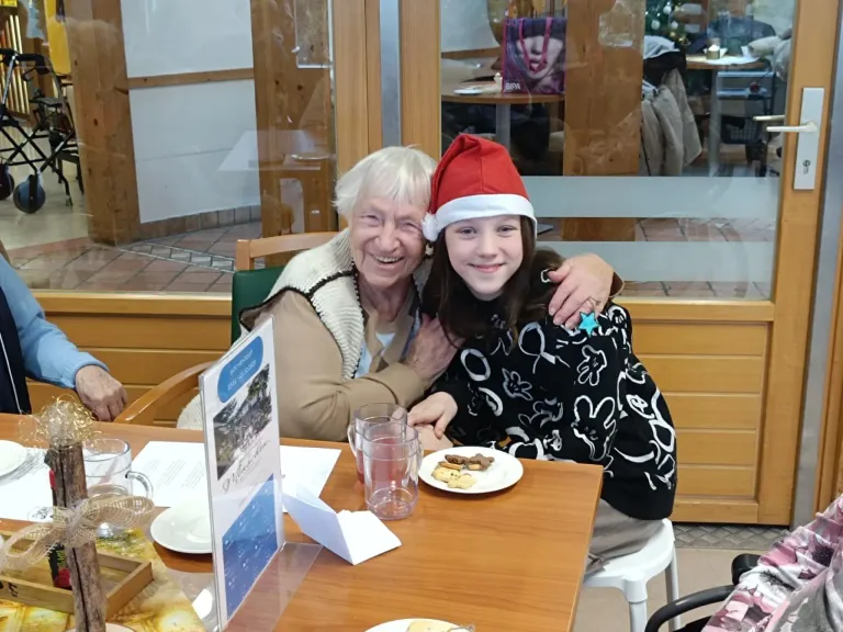 Eine ältere Frau und ein junges Mädchen sitzen lächelnd am Tisch. Das Mädchen trägt einen Weihnachtsmannhut und sie teilen sich einen Teller mit Keksen. Hinter ihnen ist eine Glaswand, die eine Restaurantumgebung zeigt.