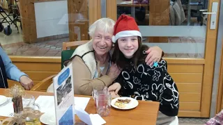 Eine ältere Frau und ein junges Mädchen sitzen lächelnd am Tisch. Das Mädchen trägt einen Weihnachtsmannhut und sie teilen sich einen Teller mit Keksen. Hinter ihnen ist eine Glaswand, die eine Restaurantumgebung zeigt.