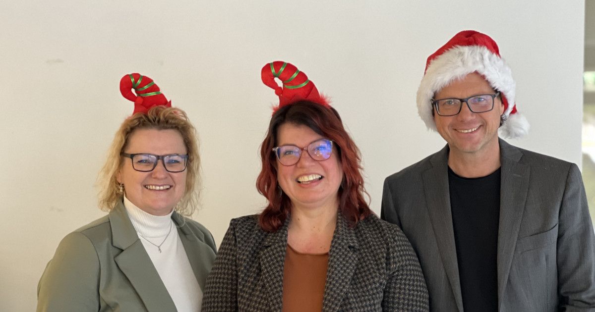 Drei Personen mit festlichen Mützen posieren für ein Foto vor einer schlichten Wand. Die linke Person trägt eine rote Elfarmband, die mittlere eine Zuckerstangenmütze und die rechte einen Weihnachtsmannhut.