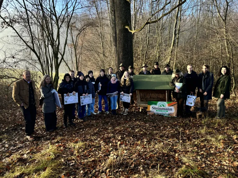 Eine Gruppe von Menschen steht in einem Wald, hält weiße Papiere in der Hand, mit einer grünen Struktur und einem Schild mit der Aufschrift Steinmann dahinter.