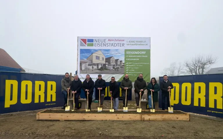 Gruppe von Menschen mit Schaufeln vor einem Baubanner, das Wohnhäuser und Text auf Deutsch zeigt.