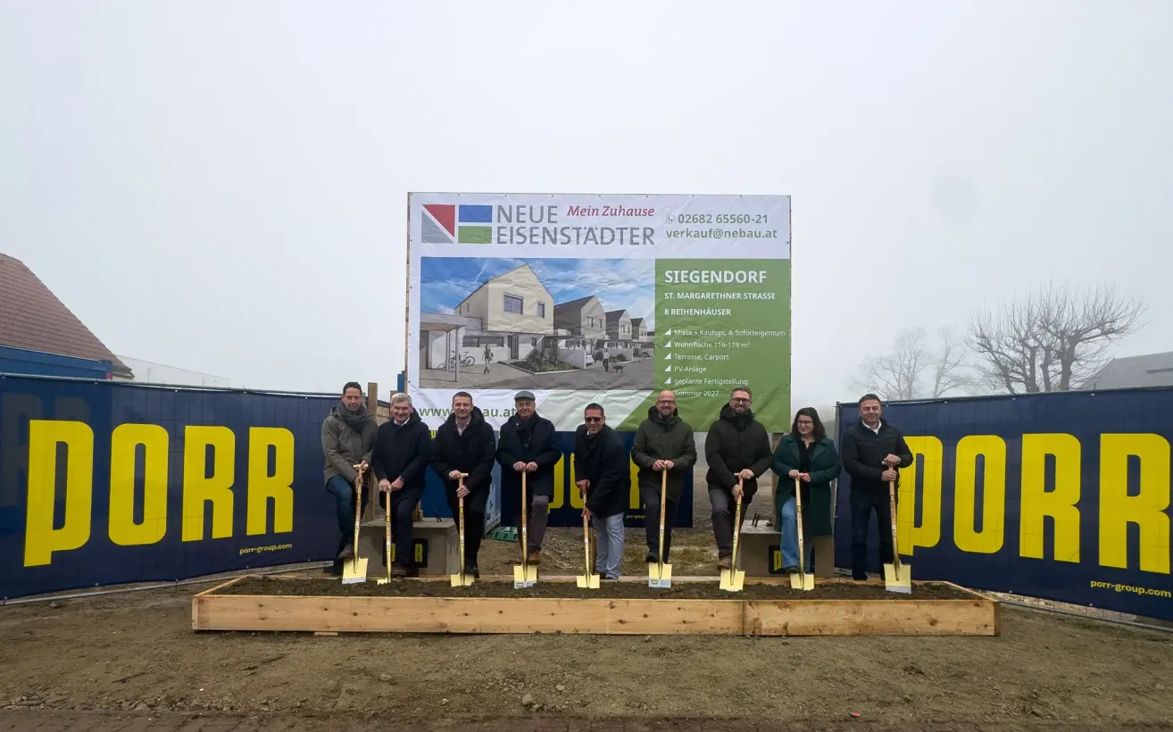 Gruppe von Menschen mit Schaufeln vor einem Baubanner, das Wohnhäuser und Text auf Deutsch zeigt.