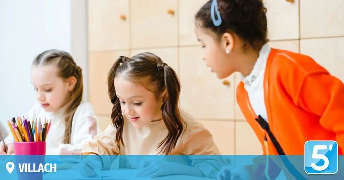 Drei Mädchen sitzen an einem Schreibtisch. Eine konzentriert sich auf ihre Arbeit, eine andere spricht mit ihr. Ein Stiftehalter steht auf dem Tisch. Das Wort Villach befindet sich in der Ecke.
