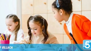 Drei Mädchen sitzen an einem Schreibtisch. Eine konzentriert sich auf ihre Arbeit, eine andere spricht mit ihr. Ein Stiftehalter steht auf dem Tisch. Das Wort Villach befindet sich in der Ecke.