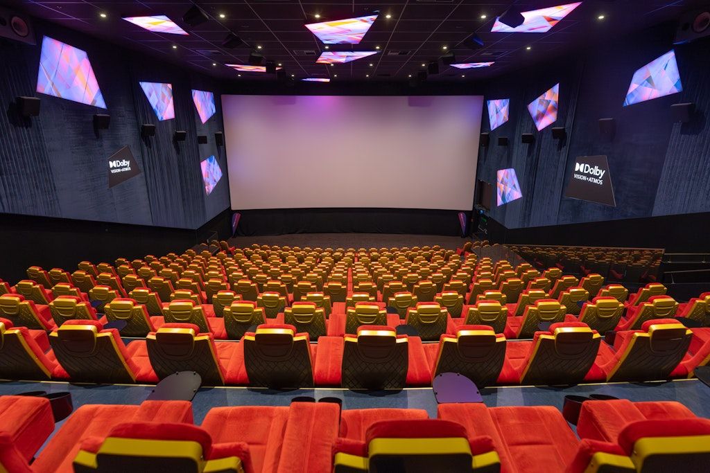 Ein leeres Kino mit einer großen Leinwand und mehreren Projektoren. Die Sitze sind rot und gelb, und der Raum ist schwach beleuchtet.