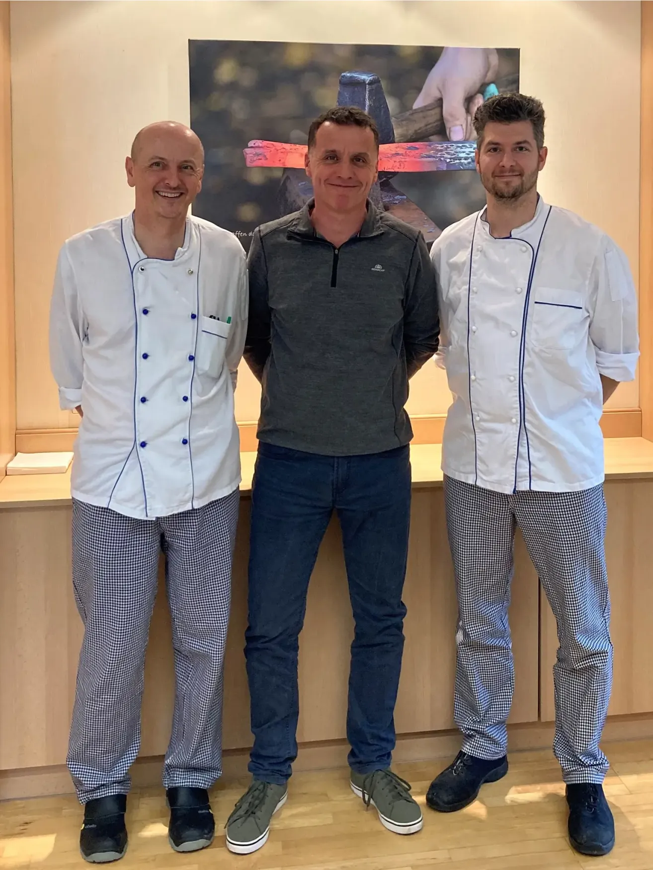 Drei Männer stehen gemeinsam vor einer Theke. Die Männer links und rechts tragen Kochuniformen, während der Mann in der Mitte eine graue Jacke trägt.