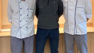 Drei Männer stehen gemeinsam vor einer Theke. Die Männer links und rechts tragen Kochuniformen, während der Mann in der Mitte eine graue Jacke trägt.