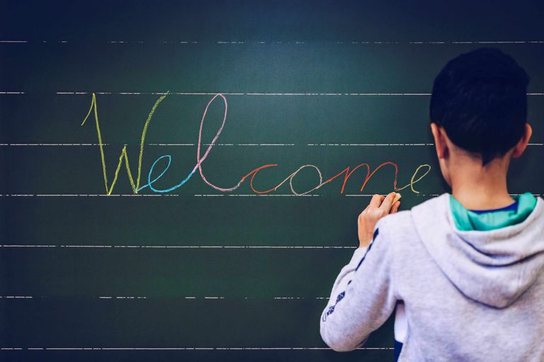 Eine Person schreibt 'Willkommen' auf eine Tafel mit Kreide in mehreren Farben. Das Brett hat liniertes Papier.