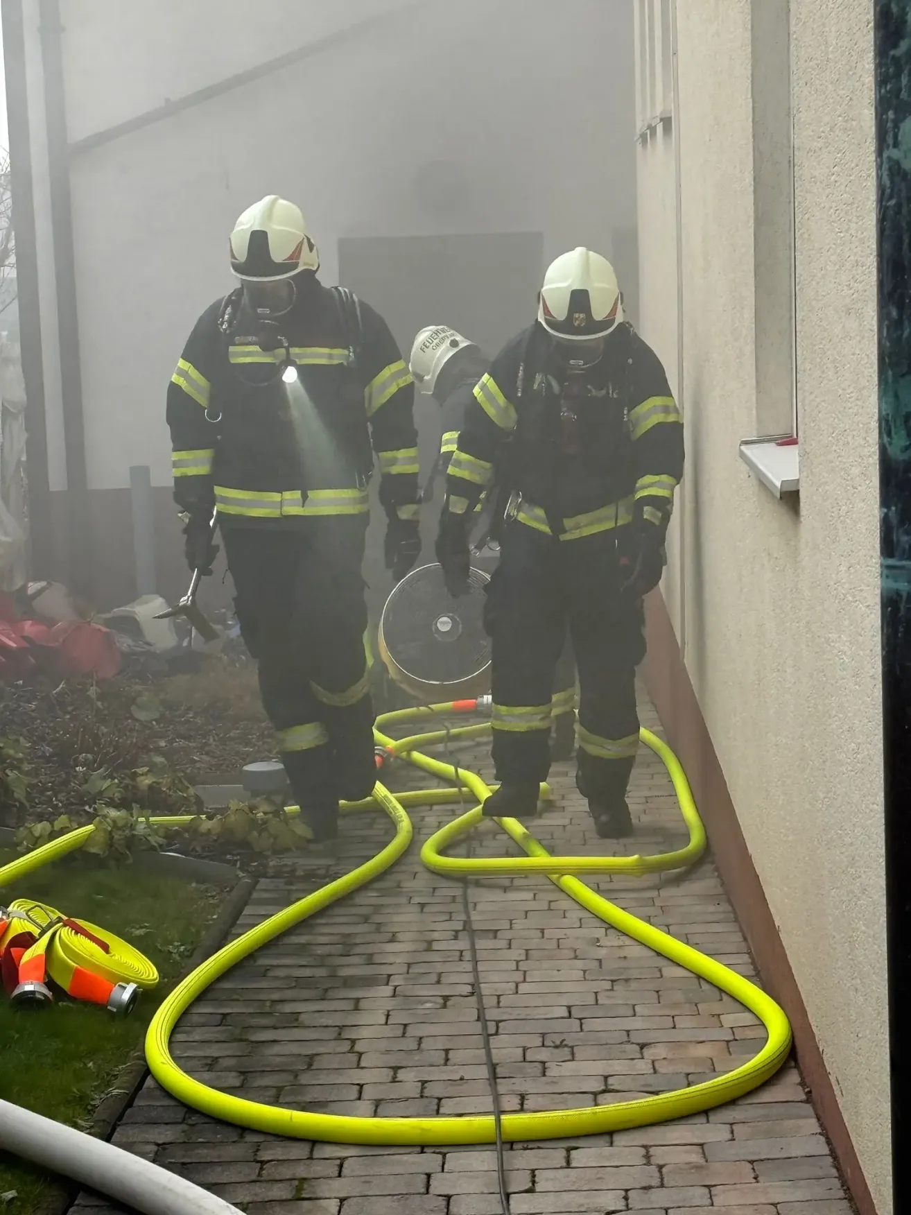 Drei Feuerwehrleute sind an einem Einsatzort tätig. Sie tragen Schutzausrüstung und Helme. Ein Feuerwehrmann hält ein Werkzeug und ein anderer steht neben einem Ventilator. Sie befinden sich auf einem gepflasterten Weg mit gelben Schläuchen ausgelegt.