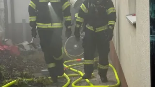 Drei Feuerwehrleute sind an einem Einsatzort tätig. Sie tragen Schutzausrüstung und Helme. Ein Feuerwehrmann hält ein Werkzeug und ein anderer steht neben einem Ventilator. Sie befinden sich auf einem gepflasterten Weg mit gelben Schläuchen ausgelegt.