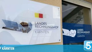 Ein weißes Schild zeigt 'Landes Rechnungshof' mit einer rot-gelben Flagge. Darunter steht 'Kärnten'. Im Hintergrund befindet sich ein Gebäude und ein blaues Schild mit dem Text 'Landes Rechnungshof'.