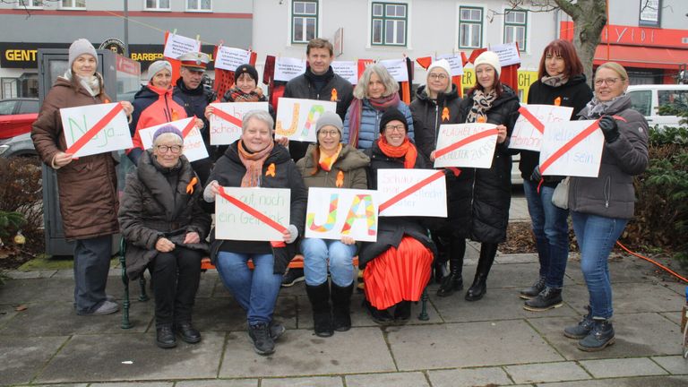 Eine Gruppe von Menschen in Winterkleidung sitzt auf einer Bank und hält Schilder mit Botschaften. Sie sind von Bannern und Schildern im Hintergrund umgeben.