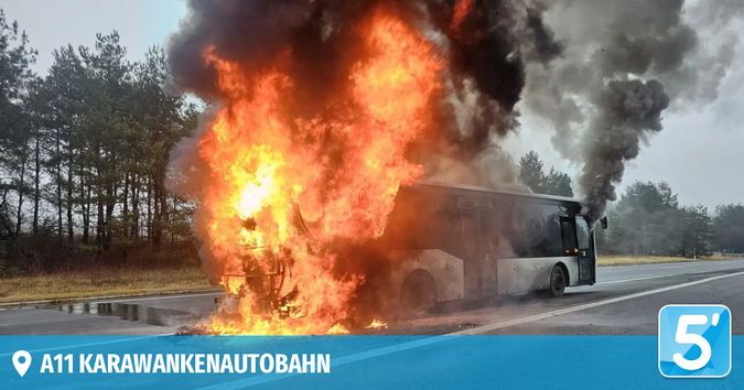 Ein brennender Bus steht am Straßenrand, mit Rauch, der herauskommt, und Bäumen im Hintergrund. Das Bild hat auch einen Wasserzeichen.