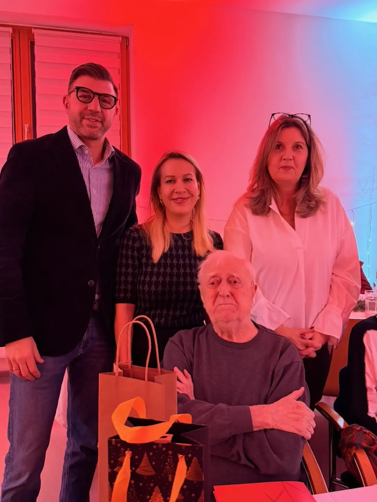 Vier Personen posieren in einem Raum mit roter und blauer Beleuchtung. Ein Mann mit Brille steht neben einer Frau mit einer Handtasche. Ein älterer Mann sitzt mit verschränkten Armen. Dahinter stehen zwei Frauen, eine mit einer Brille auf dem Kopf.