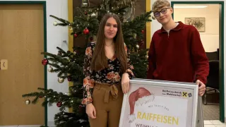 Eine Frau und ein Mann stehen neben einem Weihnachtsbaum und lächeln. Die Frau trägt ein Blumenhemd und braune Hosen. Der Mann, der eine Brille und einen roten Pullover trägt, hält ein Schild für das Raiffeisen-Weihnachtsnachtfest.