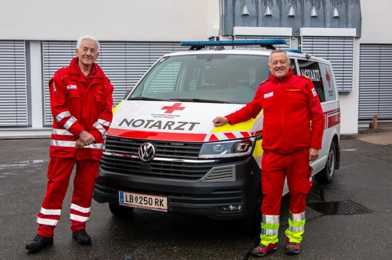Zwei Männer in roten Uniformen posieren vor einem Krankenwagen mit einem roten Kreuz auf der Seite.