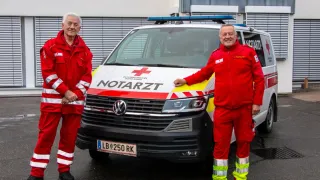 Zwei Männer in roten Uniformen posieren vor einem Krankenwagen mit einem roten Kreuz auf der Seite.