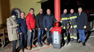 Eine Gruppe von Männern, einige in Jacken und mit Brillen, stehen um einen roten Mülleimer. Ein Mann hält einen roten Deckel. Sie alle lächeln und scheinen für ein Foto zu posieren. Hinter ihnen steht ein Gebäude mit einer gelben Wand.