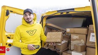 Ein Mann in einer gelben Kapuzenjacke steht neben einem gelben Van, hält eine Box mit Punkten und lächelt in die Kamera, umgeben von vielen Kisten im Van.