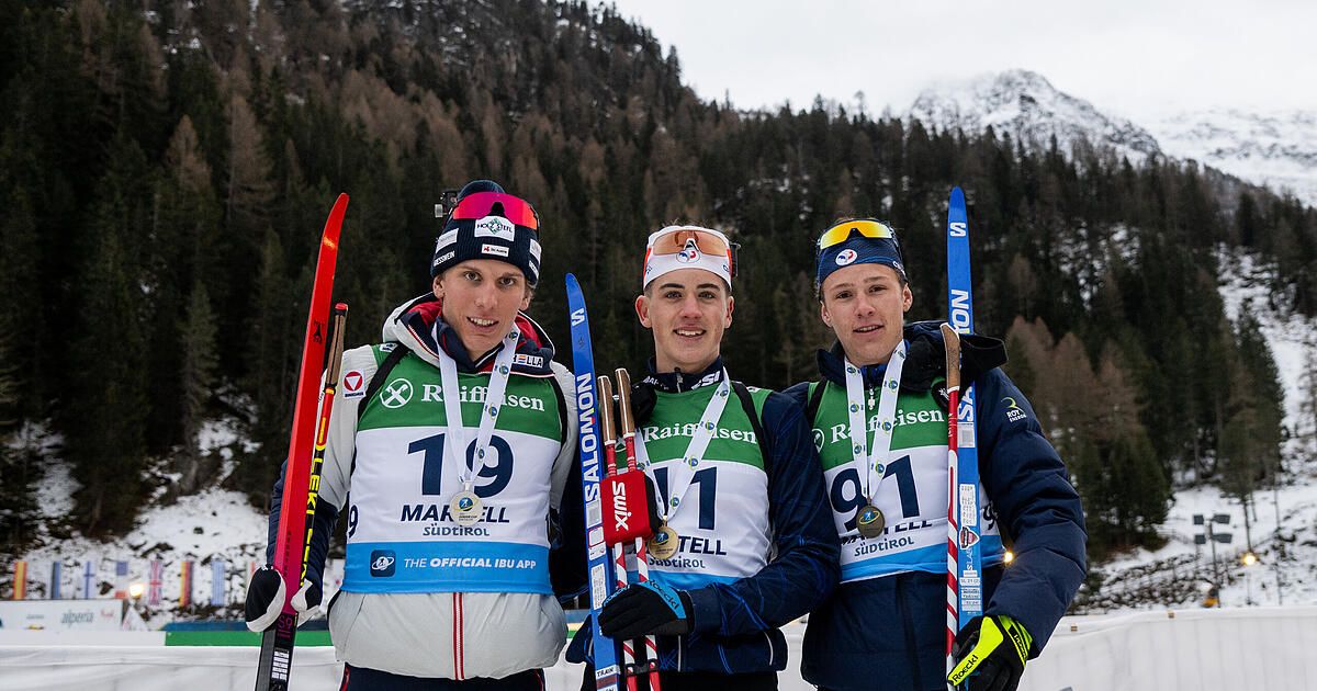Drei Skifahrer stehen auf einem verschneiten Berg, jeder mit Medaillen um den Hals. Der Skifahrer links hat die Nummer 19, der in der Mitte die Nummer 11 und der rechts die Nummer 91. Sie halten Skistöcke.