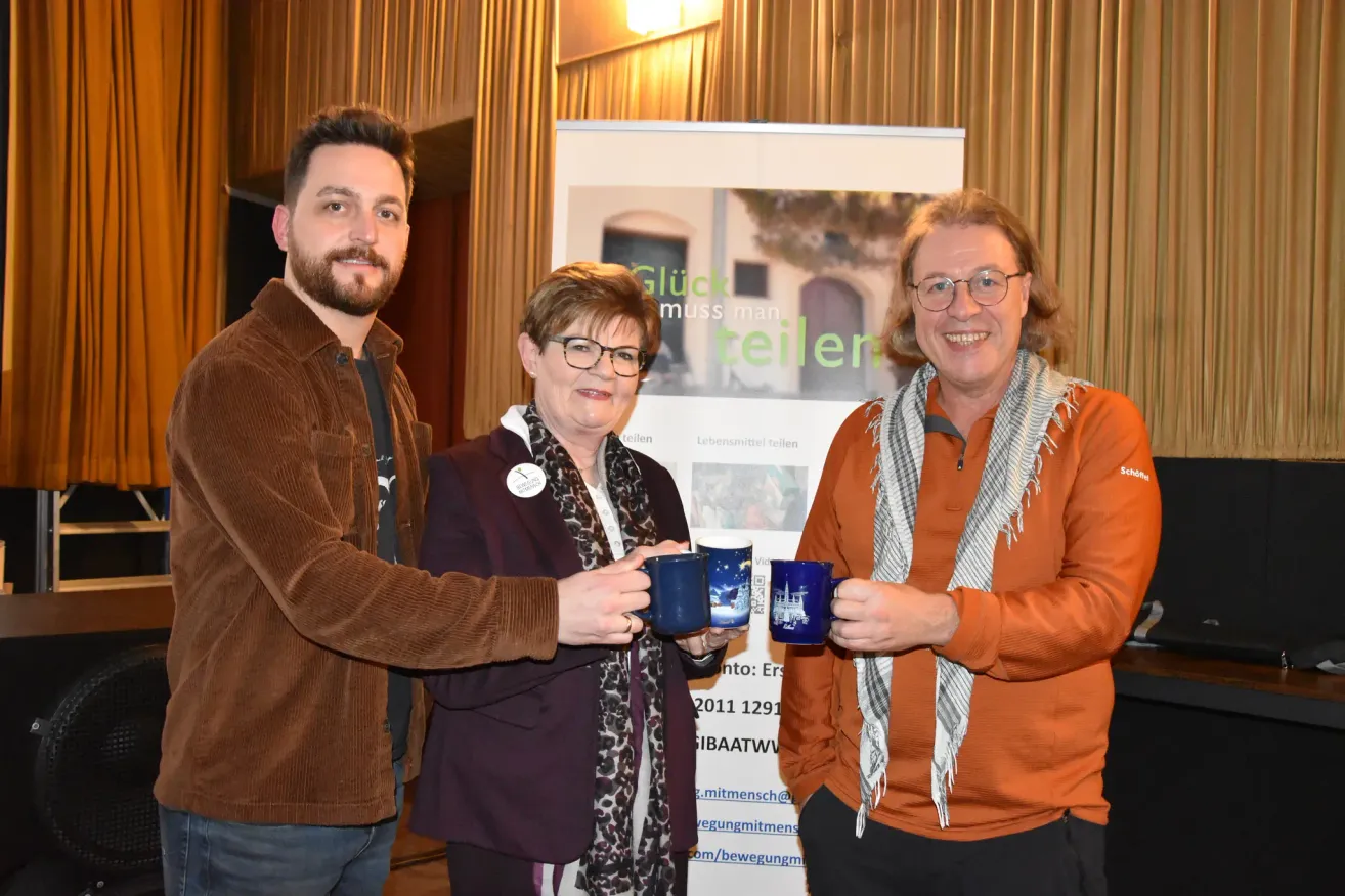 Drei Personen stehen zusammen, lächeln und halten blaue Tassen. Eine Frau in der Mitte trägt eine Brille und einen Schal. Hinter ihnen befindet sich ein Banner mit Text und einem Bild.