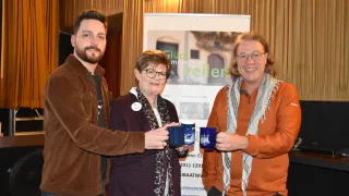 Drei Personen stehen zusammen, lächeln und halten blaue Tassen. Eine Frau in der Mitte trägt eine Brille und einen Schal. Hinter ihnen befindet sich ein Banner mit Text und einem Bild.