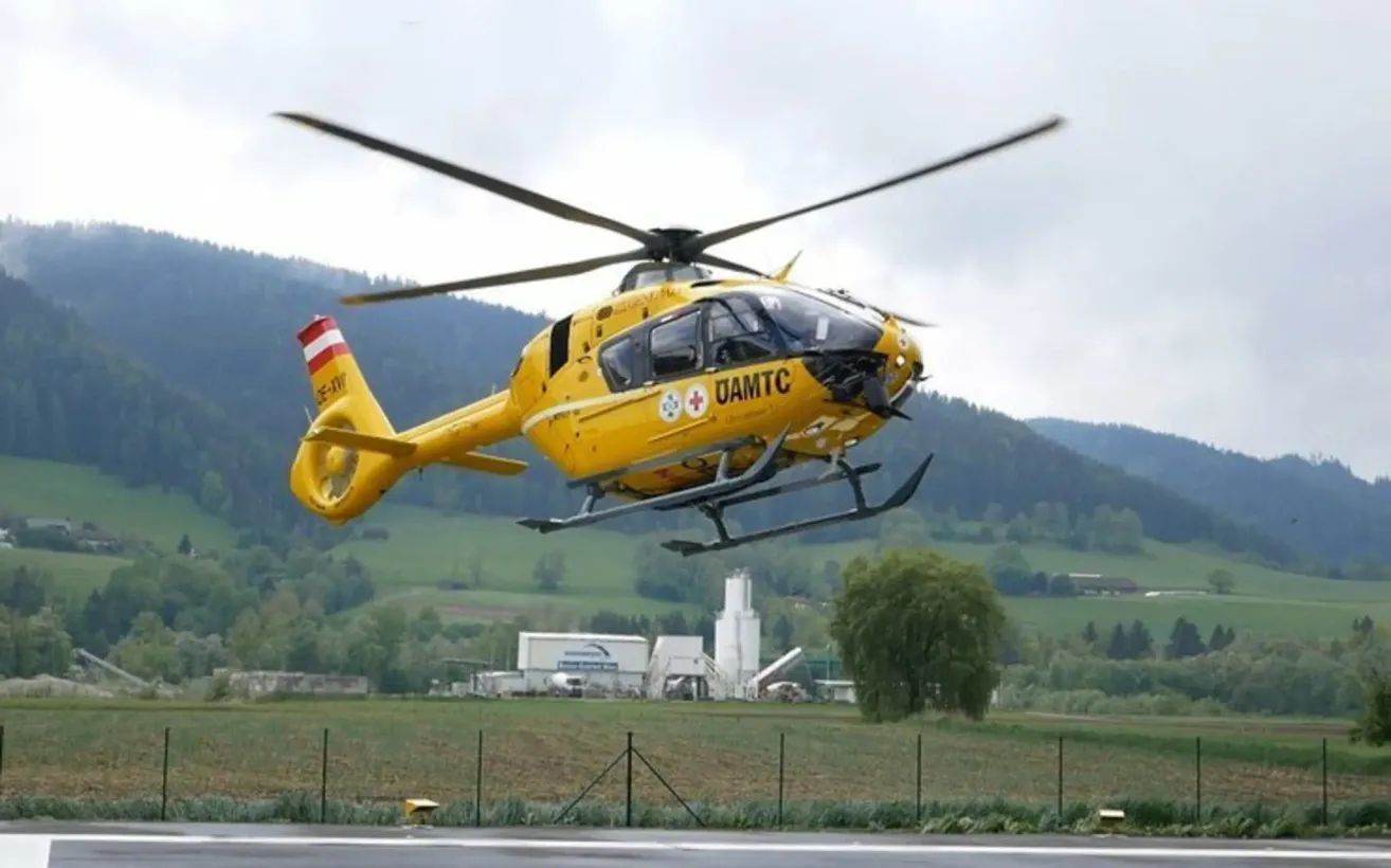 Ein gelber Hubschrauber mit der Aufschrift UAMTC fliegt über einem Feld mit Zaun und Bäumen. Berge sind im Hintergrund zu sehen.
