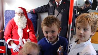 Der Weihnachtsmann in rotem Gewand steht neben einem lächelnden Jungen in einem Bus, der Schaffner steht hinter ihnen.
