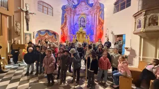 Eine Gruppe von Kindern ist in einer Kirche versammelt, einige halten Stöcke. Sie stehen vor einem Altar mit einem Gemälde, und ein Erwachsener steht hinter ihnen.