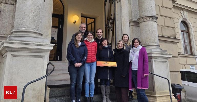 Eine Gruppe von Menschen steht auf den Stufen eines Gebäudes mit Säulen. Eine Frau hält ein gelbes Schild mit der Aufschrift 'Österreichisches Vulkanland.' Die Menschen sind für den Winter gekleidet.