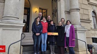 Eine Gruppe von Menschen steht auf den Stufen eines Gebäudes mit Säulen. Eine Frau hält ein gelbes Schild mit der Aufschrift 'Österreichisches Vulkanland.' Die Menschen sind für den Winter gekleidet.