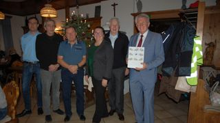 Fünf Personen stehen zusammen in einem Raum. Ein Mann hält ein Zertifikat und lächelt. Ein Weihnachtsbaum steht hinter ihnen.