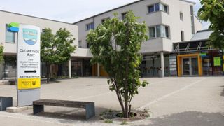 Ein modernes Gebäude mit mehreren Fenstern steht hinter einem kleinen Baum auf einem Innenhof. Eine Bank ist in der Nähe platziert, und es gibt mehr Bäume und Pflanzen im Bereich.