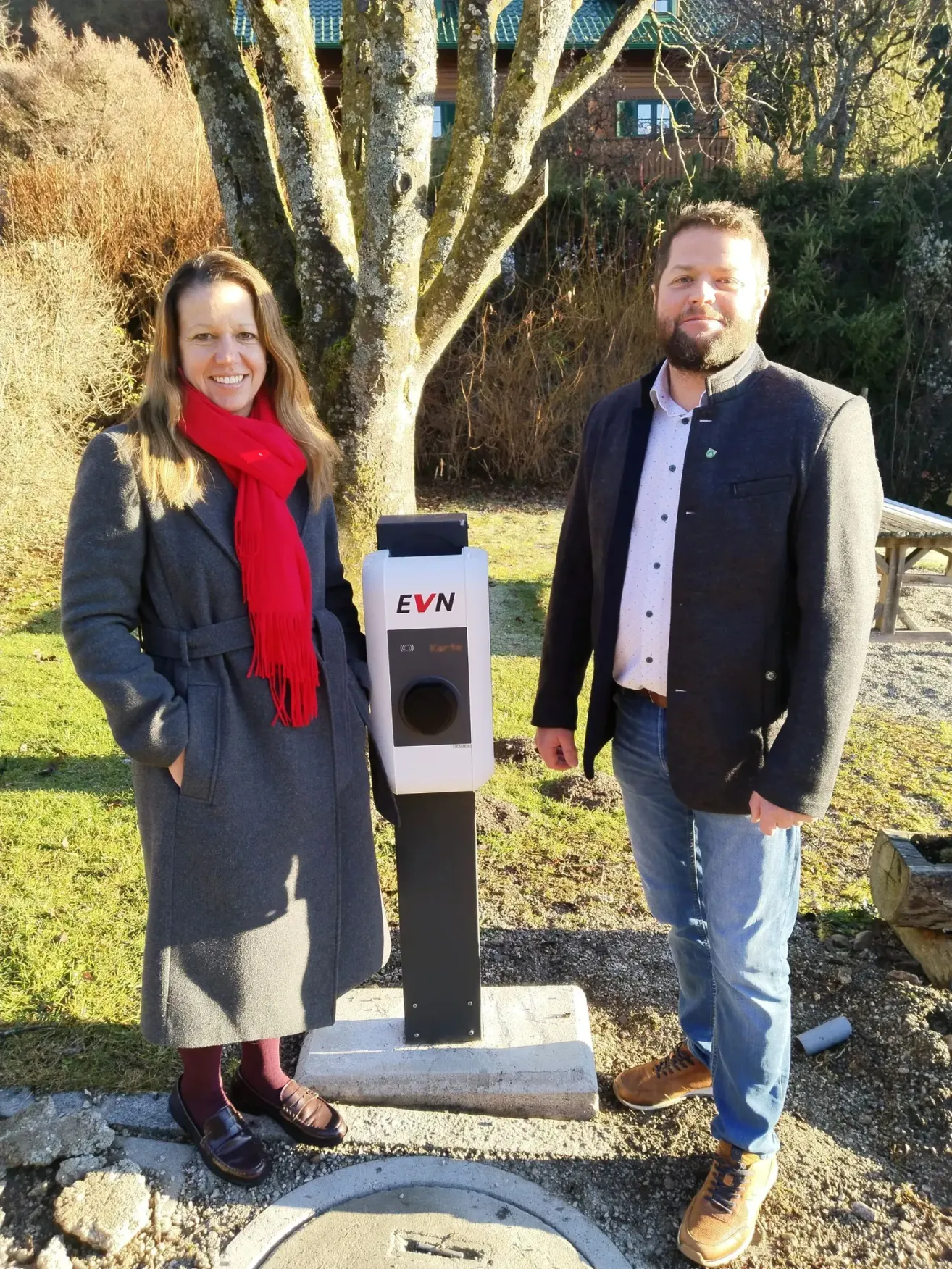 Eine Frau in einem grauen Mantel und ein Mann in einem schwarzen Mantel und Jeans stehen neben einer weißen EVN-Ladestation.