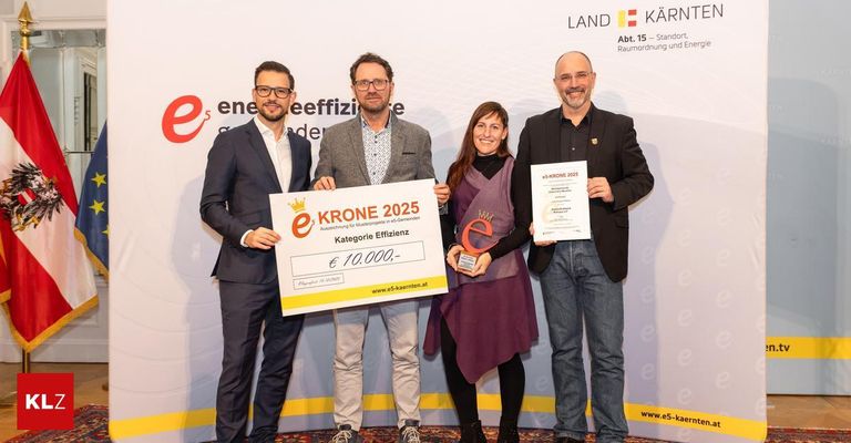 Vier Personen stehen und halten ein großes gelbes Zertifikat, eine Trophäe und ein Stück Papier. Die Frau hält eine Trophäe und ein Papier, während die beiden Männer ein Zertifikat und ein Papier halten. Sie tragen alle formelle Kleidung und scheinen sich bei einer Preisverleihung zu befinden.