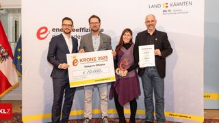 Vier Personen stehen und halten ein großes gelbes Zertifikat, eine Trophäe und ein Stück Papier. Die Frau hält eine Trophäe und ein Papier, während die beiden Männer ein Zertifikat und ein Papier halten. Sie tragen alle formelle Kleidung und scheinen sich bei einer Preisverleihung zu befinden.