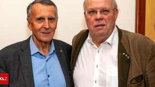 Zwei Männer stehen in einem Studio nebeneinander. Einer trägt einen grauen Mantel und blaues Hemd, der andere einen braunen Mantel und weißes Hemd.