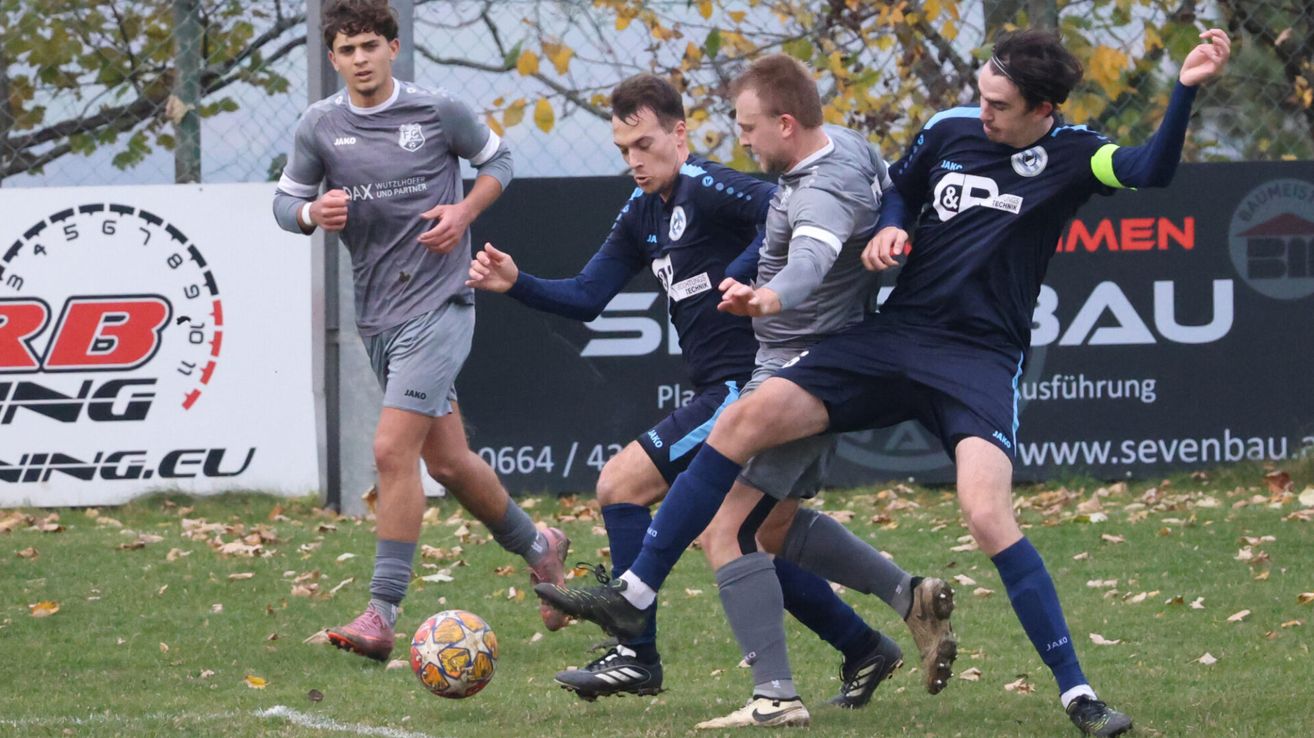 Vier Fußballspieler sind auf dem Feld und tragen Trikots. Sie kämpfen um den Ball, wobei einer der Spieler kurz davor ist, ihn zu schießen. Das Feld hat eine weiße Linie und ist von einem Zaun umgeben.