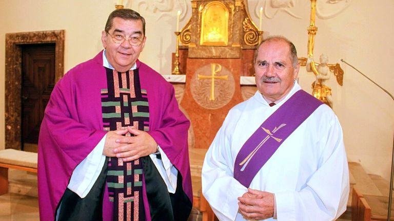 Zwei Priester stehen vor einem Kirchenaltar, einer in Lila und der andere in Weiß. Hinter ihnen befindet sich ein goldenes Kreuz und zwei Kerzen.