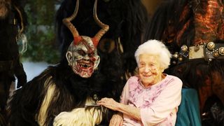 Eine ältere Frau sitzt neben einer Person, die eine gehörnte, teuflische Maske trägt. Die Frau lächelt, während die Person in der Maske Blut um den Mund hat.