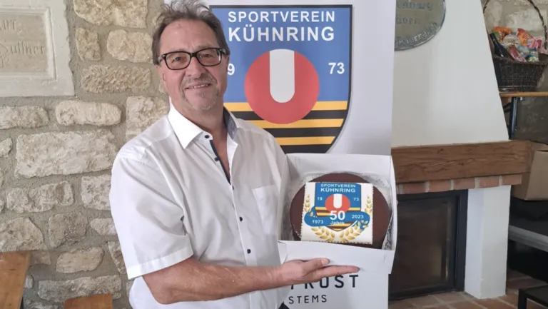 Ein Mann in einem weißen Hemd hält eine Kuchenbox mit einem Kuchen darin, der 50 Jahre Mitgliedschaft beim Sportverein Kühnring feiert.