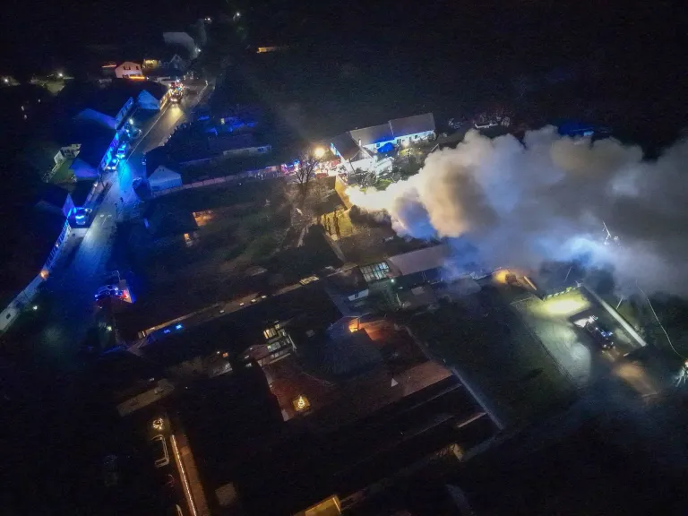 Eine Luftaufnahme einer Stadt bei Nacht zeigt ein großes Gebäude mit Rauch. Notfallleuchten blinken, und ein Feuerwehrwagen ist sichtbar.