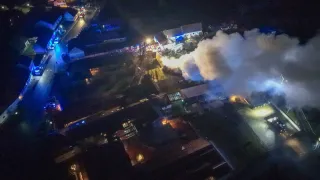 Eine Luftaufnahme einer Stadt bei Nacht zeigt ein großes Gebäude mit Rauch. Notfallleuchten blinken, und ein Feuerwehrwagen ist sichtbar.