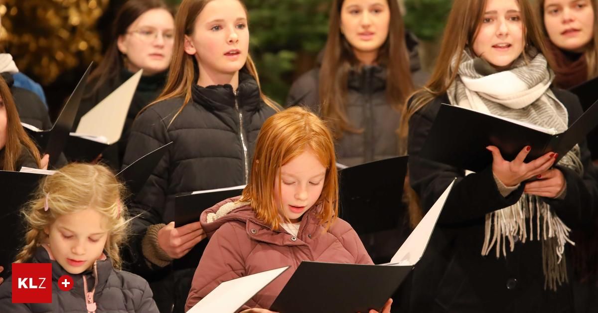 Ein Chor junger Mädchen, einige mit Brille und Schals, halten Notenblätter und singen in einer Freiluftumgebung.